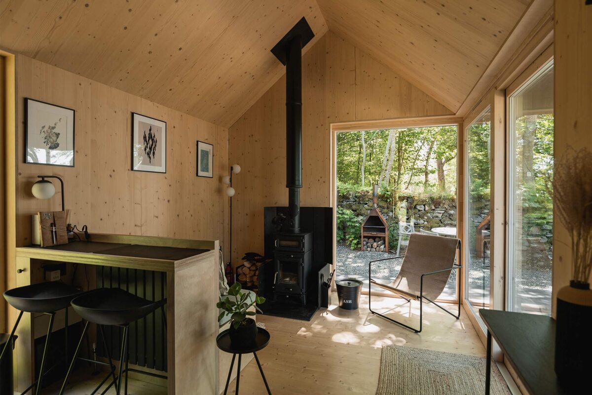 A wood burning stove stands before glazed doors opening onto a stone-walled patio with a pizza oven, framed prints on the timber wall and stools at a breakfast bar.